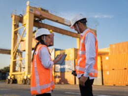 Two workers in hard hats discuss plans near shipping containers.