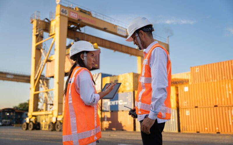 Two workers in hard hats discuss plans near shipping containers.