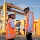 Two workers in hard hats discuss plans near shipping containers.