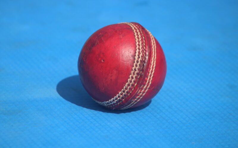 A red cricket ball sits on a blue surface.