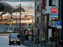 A car driving on a city street past a building with a large dinosaur mural