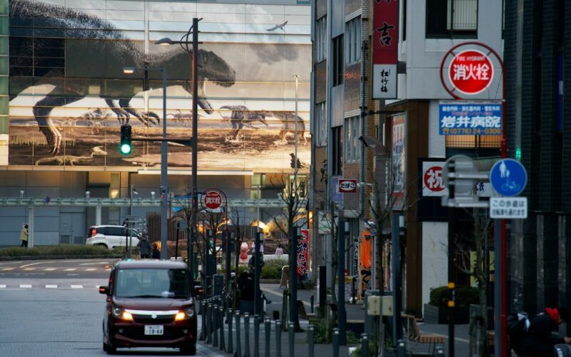 A car driving on a city street past a building with a large dinosaur mural