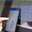 A hand pointing at a smartphone displaying currency quotes against a laptop