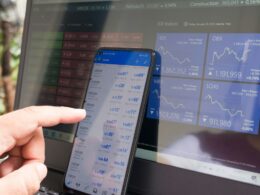 A hand pointing at a smartphone displaying currency quotes against a laptop