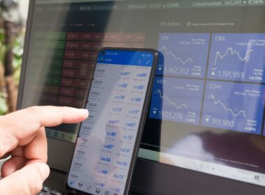 A hand pointing at a smartphone displaying currency quotes against a laptop
