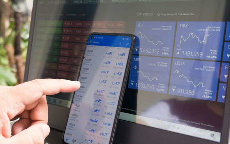 A hand pointing at a smartphone displaying currency quotes against a laptop